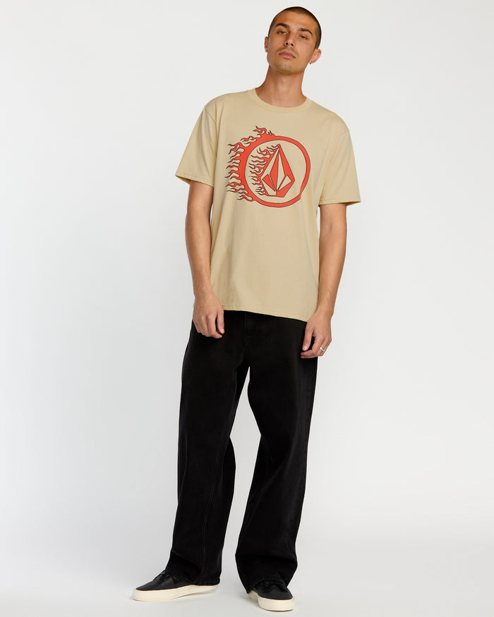A man wears the Volcom Men's Circle Stone Short Sleeve Tee in beige, featuring a red graphic, with black pants and shoes, standing against a plain white background.
