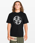 A person with long, wavy brown hair faces the camera wearing Volcom’s Men's Circle Stone Split Short Sleeve Tee in black, featuring a large white geometric graphic, paired with olive green shorts.