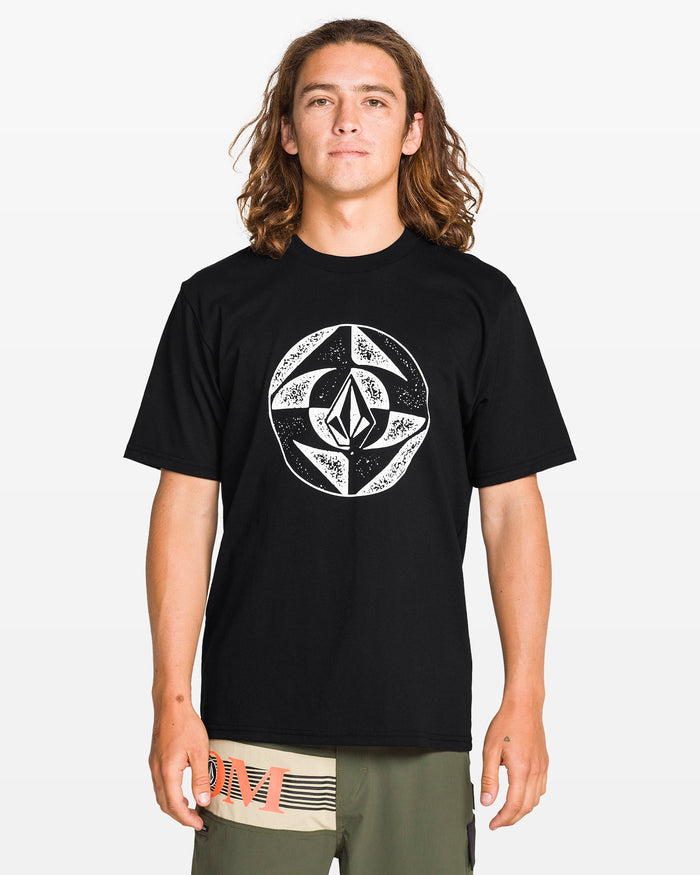 A person with long, wavy brown hair faces the camera wearing Volcom’s Men's Circle Stone Split Short Sleeve Tee in black, featuring a large white geometric graphic, paired with olive green shorts.