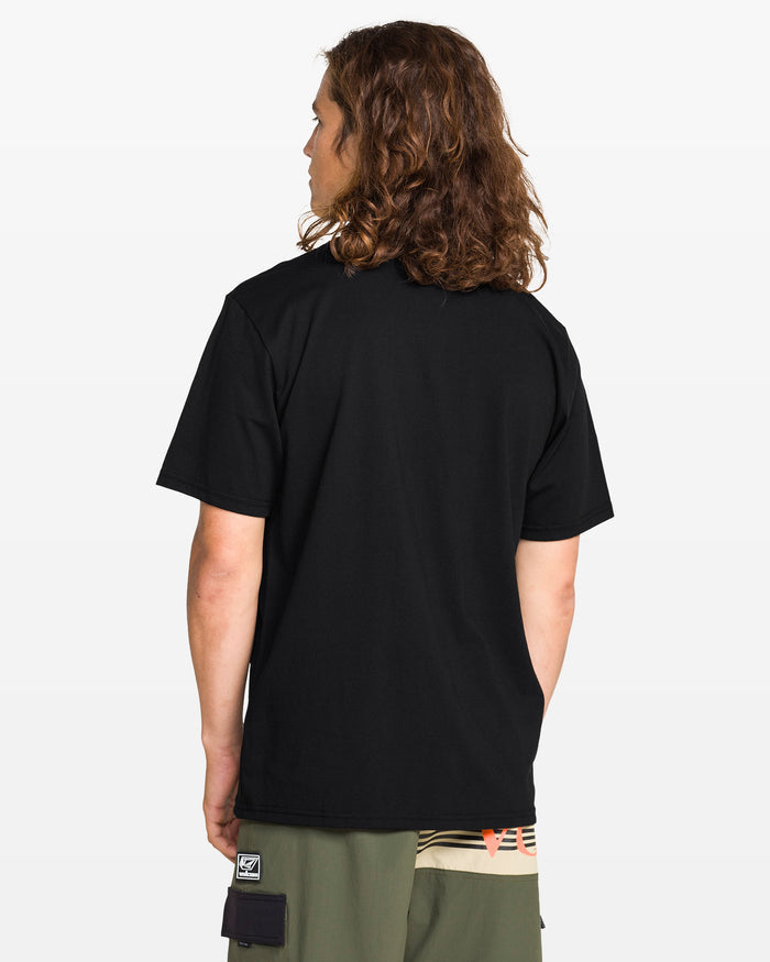 A person with long, wavy brown hair stands with their back to the camera, wearing a Volcom Men's Circle Stone Split Short Sleeve Tee in black and green shorts with multicolored accents and a Circle Stone patch. The background is white.