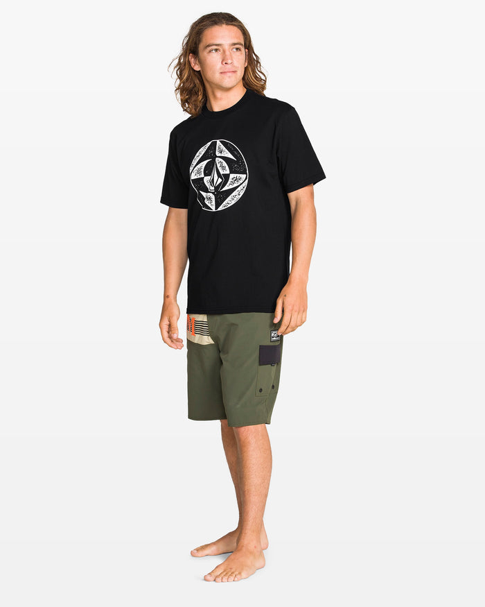 A young man with long hair stands barefoot against a white background, wearing the Volcom Men's Circle Stone Split Short Sleeve Tee and olive green board shorts with patchwork designs.