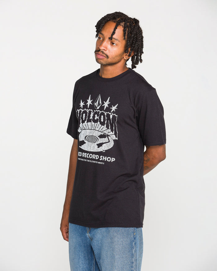 A man with short dreadlocks is wearing the Volcom Men's Collector Short Sleeve Tee in washed black, featuring crackle-ink "POISON RECORD SHOP" artwork, paired with blue jeans. He stands against a plain white background with his hands behind his back, looking slightly to the side.