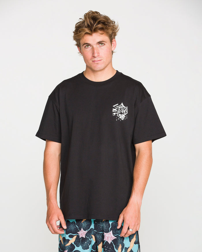 A young man with tousled hair stands against a plain white background, looking at the camera with a neutral expression. He is wearing the Volcom Men's Concrete Jungle Short Sleeve Tee, crafted from heavyweight ringspun cotton, paired with patterned shorts. The crew neck t-shirt features a bold graphic that adds an urban edge to his relaxed look.