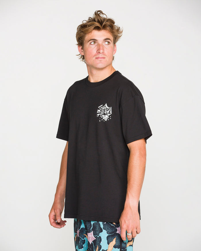 A young man with tousled hair wears the Volcom Men's Concrete Jungle Short Sleeve Tee in black, a heavyweight ringspun cotton shirt featuring a small white graphic on the left chest. He pairs it with colorful patterned shorts, and the classic crew neck tee stands out against a plain white background as he looks slightly to the side.