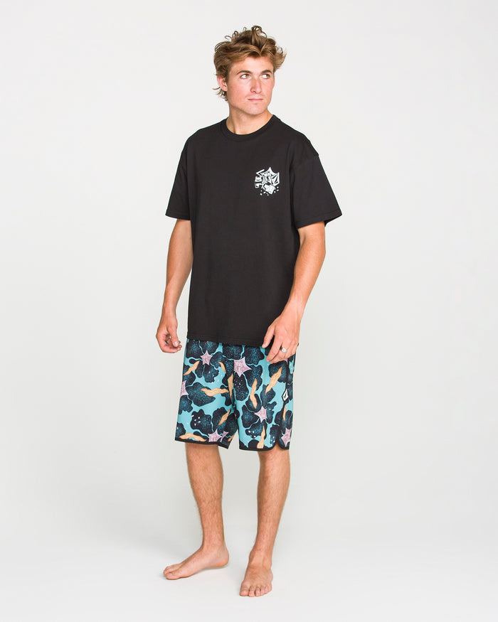 A barefoot person stands on a plain background, wearing the Volcom Men's Concrete Jungle Short Sleeve Tee—a black crew neck made from heavyweight ringspun cotton with a small white chest graphic—paired with blue patterned shorts.