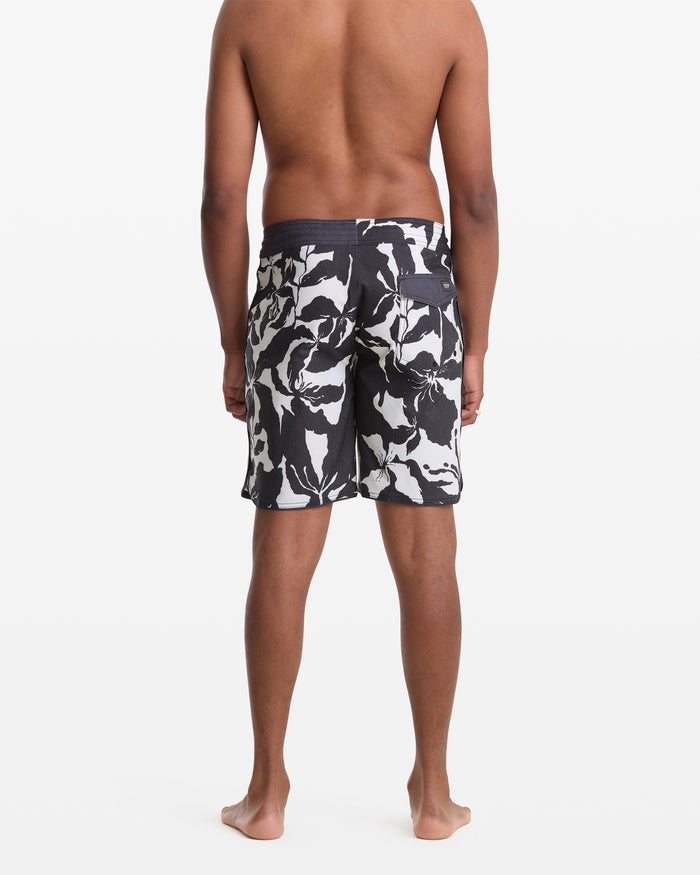 A man stands barefoot facing away, wearing Volcom Men's Contact Scallop Stoney 19 swim trunks with a black and white floral pattern, set against a plain white background.