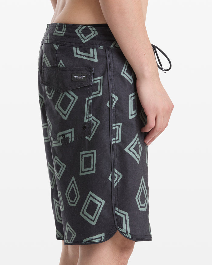 A man wearing Volcom Men's Contact Scallop Stoney 19 black shorts with a green geometric pattern and a back pocket featuring the Volcom logo patch, shown from the side against a plain white background.