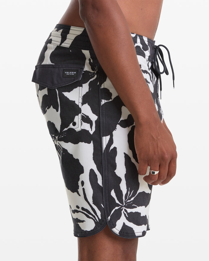 A person stands sideways wearing Volcom Men's Contact Scallop Stoney 19 board shorts with a black and white floral pattern, side pocket, and drawstring waist. Their hand is by their thigh, displaying a rectangular silver ring.