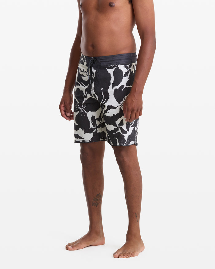 A man stands barefoot against a white background wearing Volcom Men's Contact Scallop Stoney 19 swim trunks; his upper body is shirtless, and his arms rest at his sides.