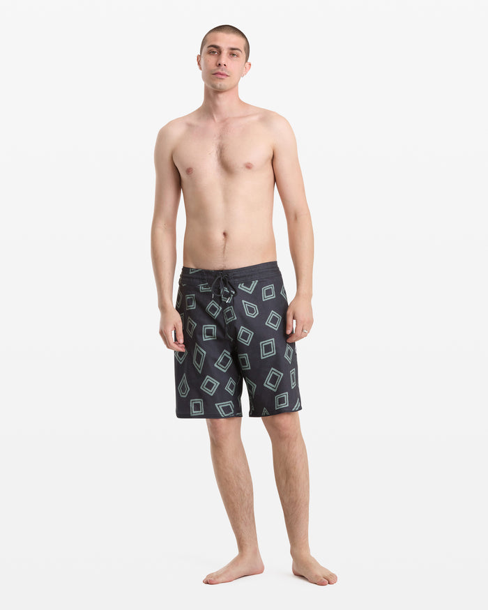 A man stands barefoot against a white background wearing Volcom Men's Contact Scallop Stoney 19 board shorts with a geometric square pattern. He is shirtless, looking at the camera in a relaxed posture.