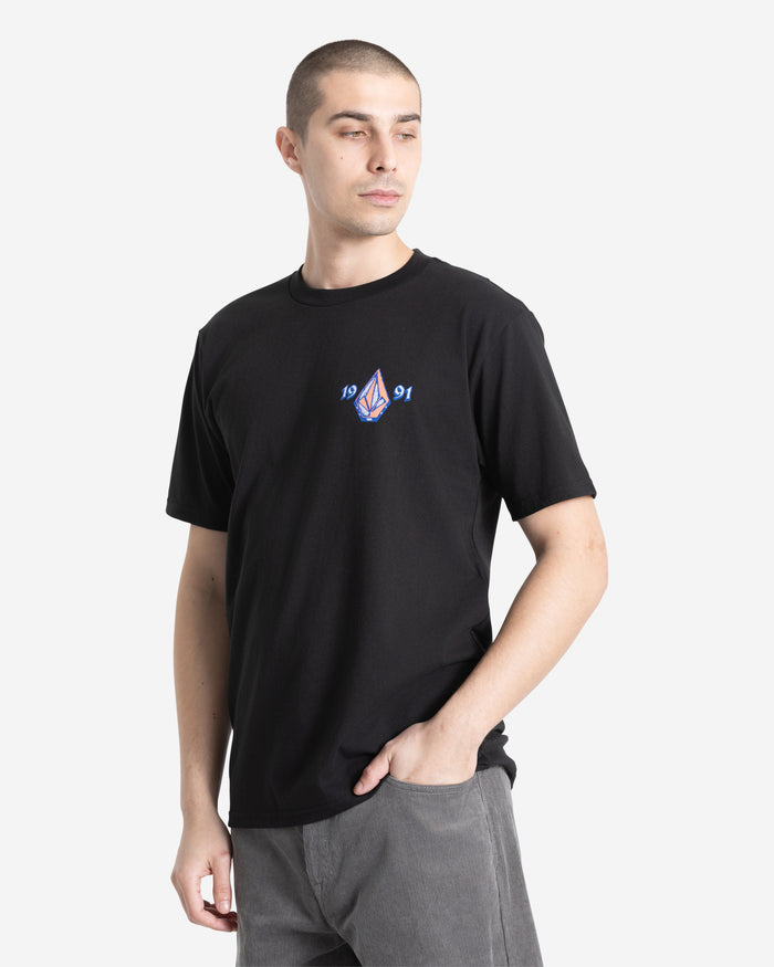 A man in a Volcom Men's Countertop Short Sleeve Tee—100% cotton, black, small chest graphic with "1991"—stands with one hand in his pocket, looking to the side against a plain white background.