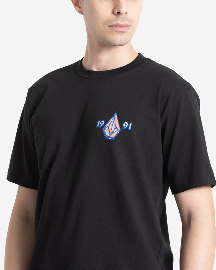 A man wears the Volcom Men's Countertop Short Sleeve Tee in black, featuring a small blue and purple geometric logo with "1991" on the chest. Made from 100% cotton, he looks slightly to the side.