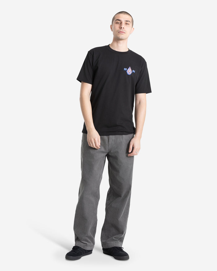 A person stands against a white background, wearing the Volcom Men's Countertop Short Sleeve Tee in black—100% cotton with a small chest graphic—paired with loose gray pants and black shoes. They have short hair and a relaxed expression, hands at their sides.