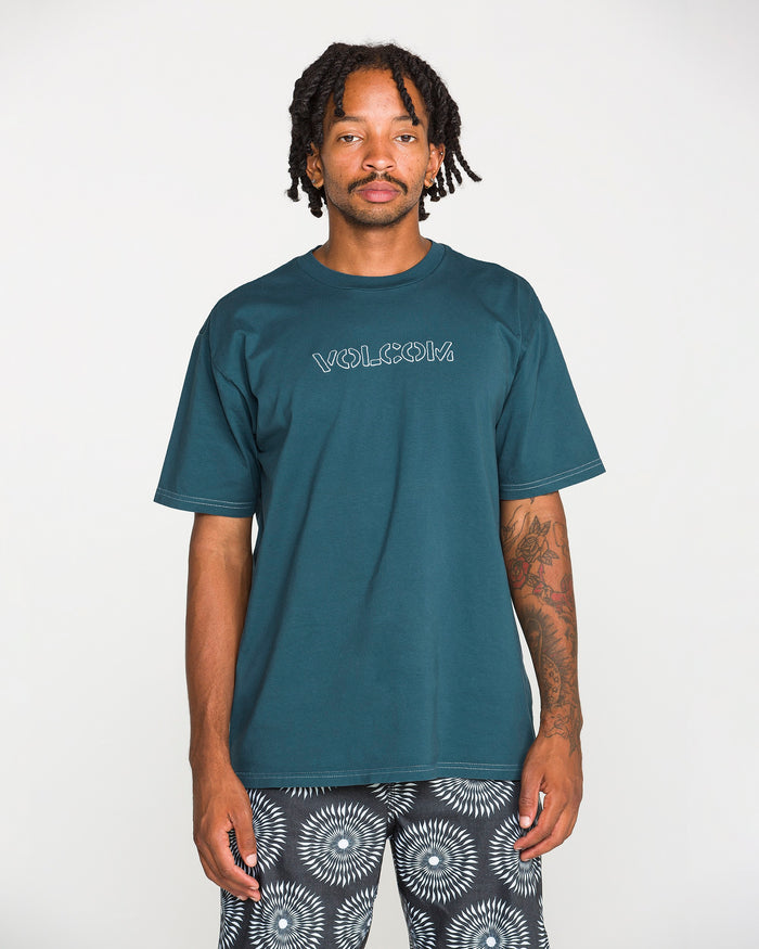 A man with short dreadlocks stands against a plain white background, looking directly at the camera, wearing the Volcom Men's Crassy Short Sleeve Tee in premium cotton with a teal graphic, paired with patterned shorts.