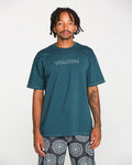 A man with short dreadlocks stands against a plain white background, looking directly at the camera, wearing the Volcom Men's Crassy Short Sleeve Tee in premium cotton with a teal graphic, paired with patterned shorts.