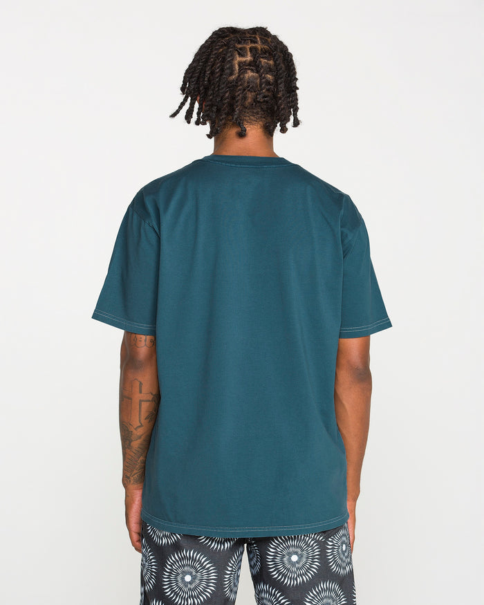 A person with short dreadlocks is standing with their back to the camera, wearing the Volcom Men's Crassy Short Sleeve Tee in teal paired with patterned black-and-white shorts. The loose fit of the t-shirt drapes comfortably, set against a plain white background.