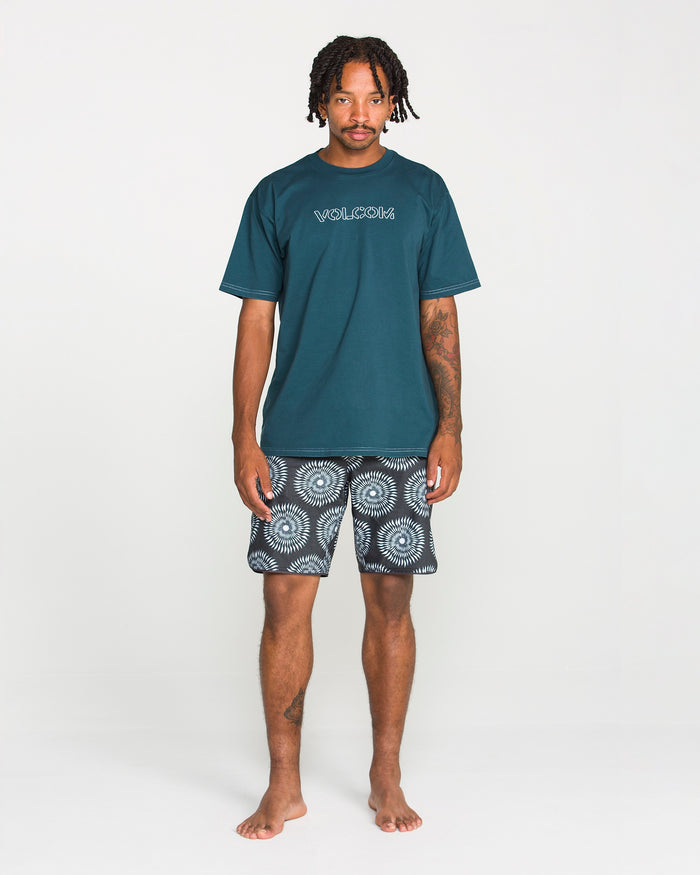 A man stands barefoot against a white background, wearing Volcom's Men's Crassy Short Sleeve Tee in teal—crafted from premium cotton with a soft-hand screenprint—and patterned black-and-white shorts.