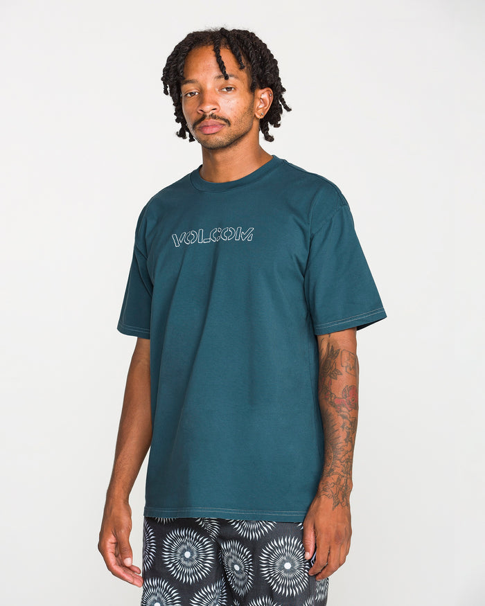 A man with short dreadlocks and arm tattoos wears the Volcom Men's Crassy Short Sleeve Tee in teal, featuring a soft-hand screenprint, paired with black-and-white patterned shorts and standing against a plain white background.