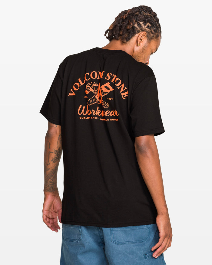 A man with dreadlocks faces away, wearing a Volcom Men's Crosscut Workwear Short Sleeve Tee in classic fit with bold orange graphics on the back, paired with blue all-cotton shorts.