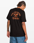 A man with dreadlocks faces away, wearing a Volcom Men's Crosscut Workwear Short Sleeve Tee in classic fit with bold orange graphics on the back, paired with blue all-cotton shorts.
