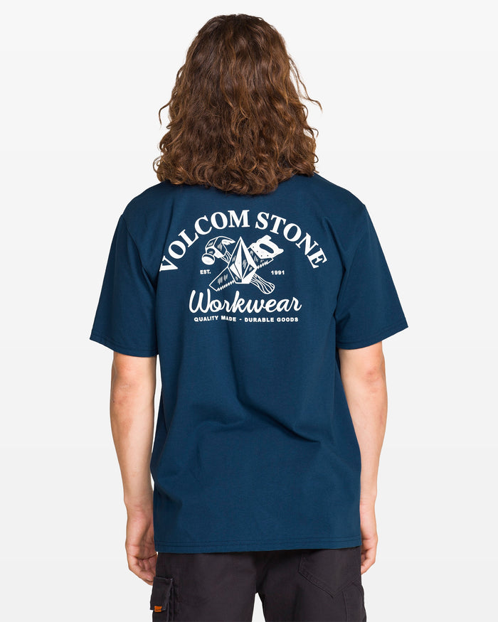 Seen from behind, a person with long, wavy hair wears a Men’s Crosscut Workwear Short Sleeve Tee by Volcom. The all-cotton shirt features a white hammer, wrench, and origami graphic on navy blue.