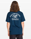 Seen from behind, a person with long, wavy hair wears a Men’s Crosscut Workwear Short Sleeve Tee by Volcom. The all-cotton shirt features a white hammer, wrench, and origami graphic on navy blue.