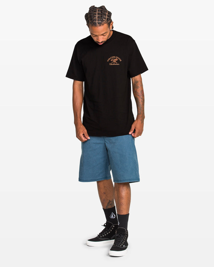 A man with braided hair looks down, wearing the Volcom Men's Crosscut Workwear Short Sleeve Tee for all-cotton comfort, paired with blue knee-length shorts, black socks with a white logo, and black sneakers against a plain white background.