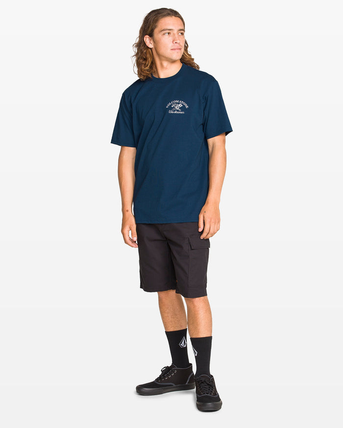 A young man with long hair wears the Volcom Men's Crosscut Workwear Short Sleeve Tee in navy, styled with black shorts, logoed black socks, and black sneakers. He stands against a plain white background, looking to his left.
