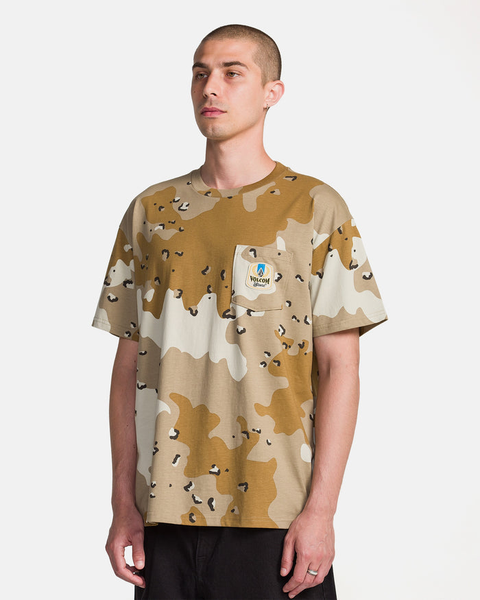 A man with short hair wears a Volcom Men's Desert Beers Pocket Short Sleeve Tee in tan and brown camo, made of heavyweight cotton. The chest pocket patch reads WGM with a cartoon tiger. He stands against a plain white background.