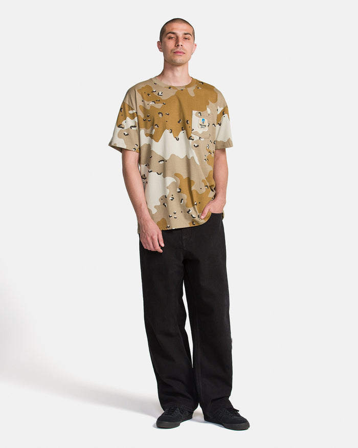A person stands against a plain white background, wearing the Volcom Men's Desert Beers Pocket Short Sleeve Tee, black pants, and black sneakers, with one hand in their pocket.