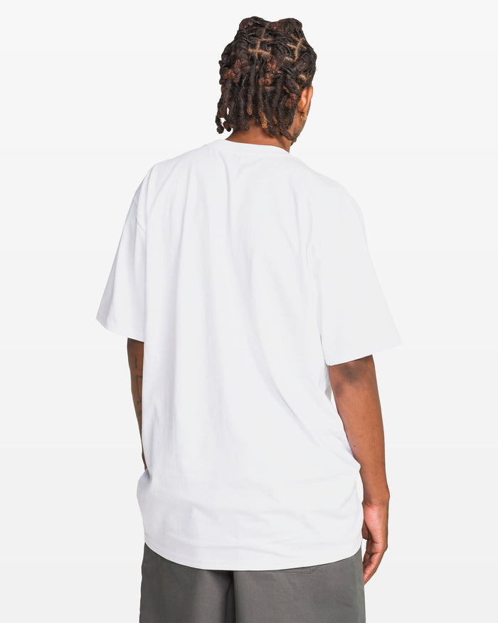 A person with short dreadlocks, seen from behind against a plain white background, wears a Volcom Men's Devilish Short Sleeve Tee in classic fit 100% cotton and dark shorts.