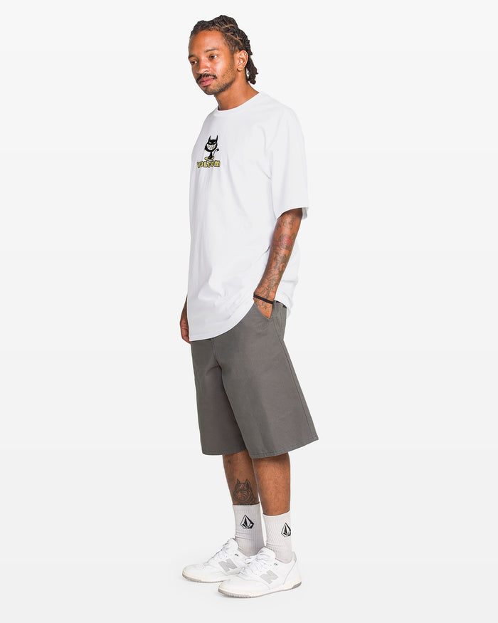 A man with braided hair stands sideways with hands in pockets, wearing a Volcom Men's Devilish Short Sleeve Tee, classic fit gray knee-length shorts, white logo socks, and white sneakers against a plain white background.