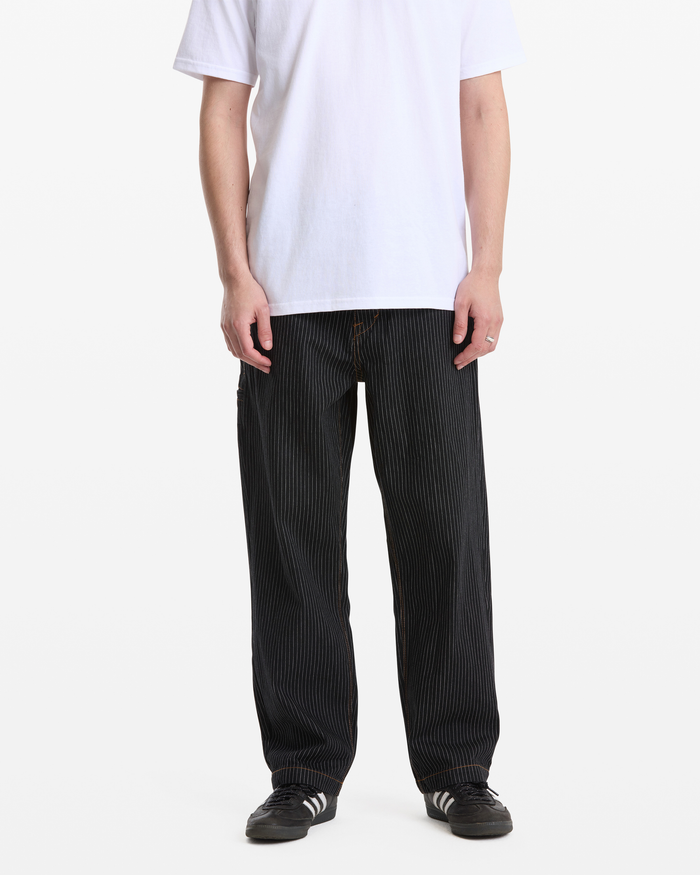 A person wearing Volcom Men's Dialed Work Pants in black pinstripes, a plain white T-shirt, and black Adidas sneakers with white stripes stands against a plain white background. The person's head is not visible.