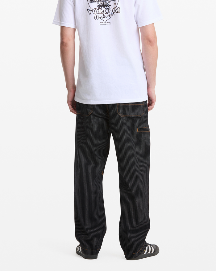 A person stands facing away against a plain white background, wearing a white Volcom T-shirt, Volcom Men's Dialed Work Pants in black pinstripe, and black and white slide sandals.