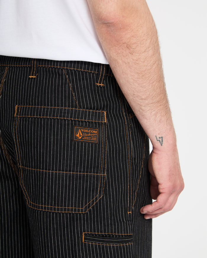 A person wears Volcom Men's Dialed Work Pants—black pinstripe with orange stitching and a brand patch on the back pocket. Their right arm, featuring a small wrist tattoo, is visible against a plain background.
