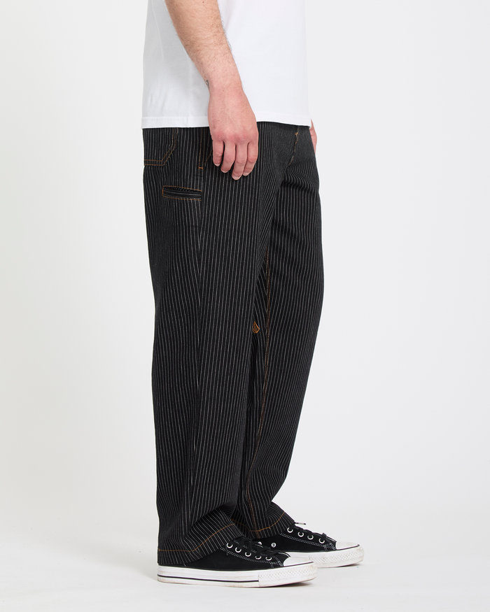 A person stands sideways against a plain white background, modeling Volcom Men's Dialed Work Pants with a white t-shirt and black low-top sneakers. Focus is on the pants.