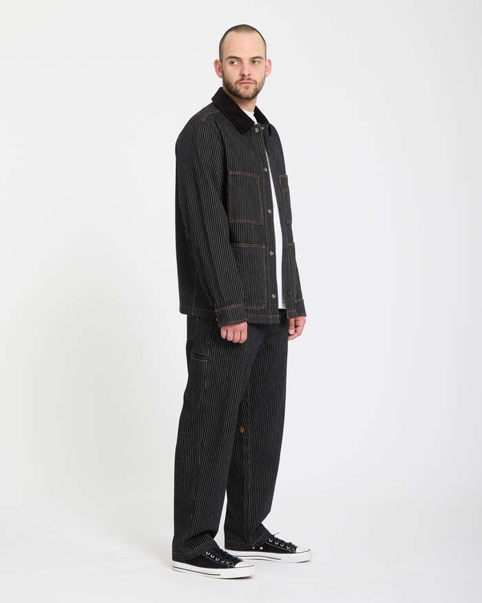 A man faces forward in a studio, wearing Volcom Men's Dialed Work Pants with orange stitching, a black pinstripe jacket, white t-shirt, and black low-top sneakers against a plain white background.