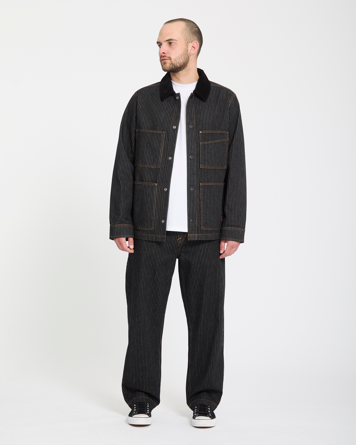 A man stands against a plain white background wearing Volcom Men's Dialed Work Pants, a dark pinstriped jacket, white t-shirt, and black sneakers. He looks slightly left with his hands by his sides.