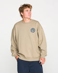 Wearing the Volcom Men's Dirty Work Crew, a loose fit khaki sweatshirt crafted from soft brushed-back fleece and detailed with a small circular black-and-white logo on the left chest, a person stands with hands in blue pants pockets, looking off to the side against a plain background.