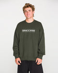 A person with short, tousled hair wears the Volcom Men's Dirty Work Crew— a green, loose fit fleece crew sweatshirt crafted from recycled polyester—paired with black pants and standing against a plain light background. The sweatshirt features white text for added style and offers both comfort and sustainability.