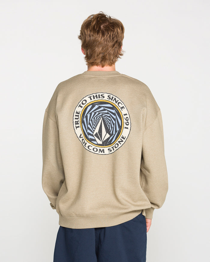 A person faces away, wearing the Volcom Men's Dirty Work Crew in a khaki tone. The sweatshirt features a loose fit and comfortable brushed-back fleece. On the back, a circular logo reading "TRUE TO THIS SINCE 1991 VOLCOM STONE" surrounds an abstract geometric design, adding distinctive style to its casual look.