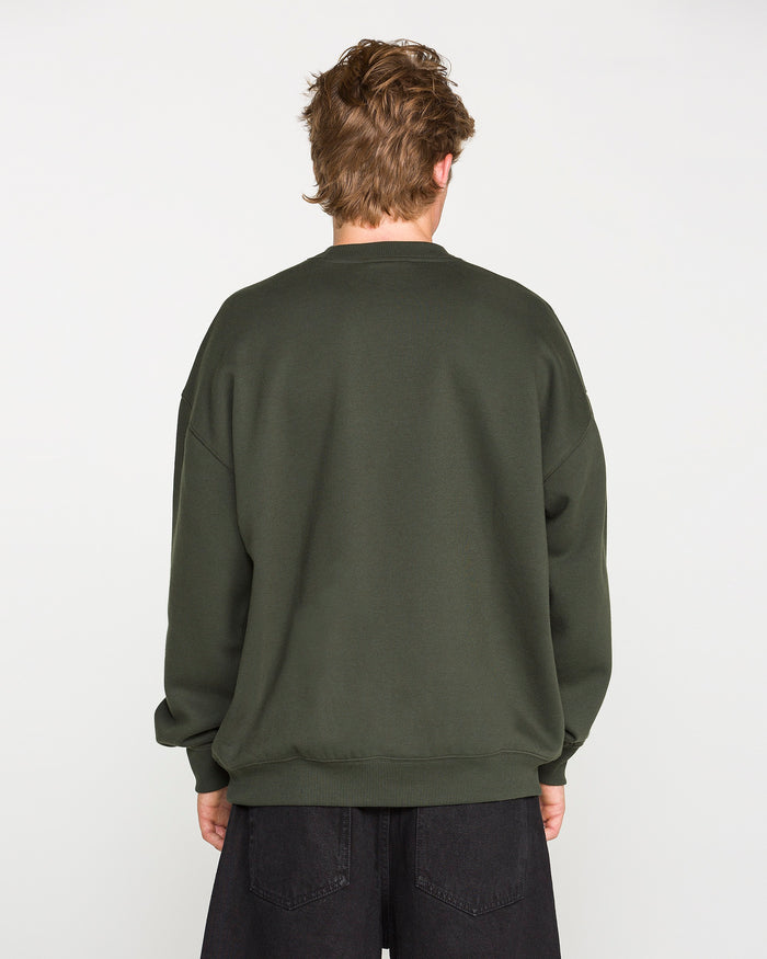 A person with short, light brown hair is facing away from the camera, wearing the Volcom Men's Dirty Work Crew—an oversized green sweatshirt crafted from recycled polyester—paired with black pants against a plain white background.