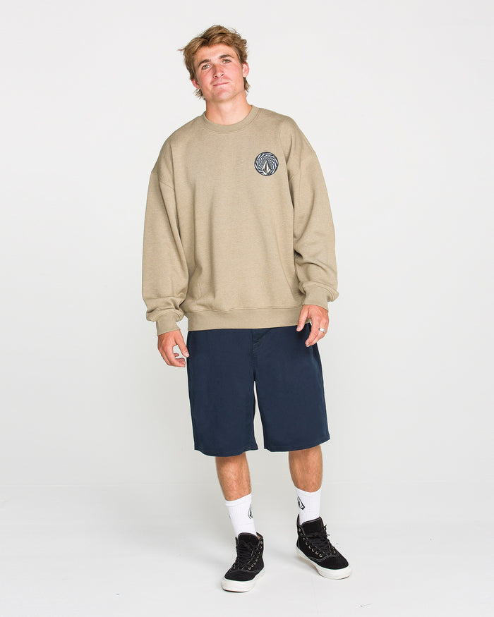 A young man stands against a plain white background, wearing the Volcom Men's Dirty Work Crew—an easygoing beige (khaki) brushed-back fleece sweatshirt with a small circular logo—paired with navy blue shorts, white socks, and black sneakers. The loose fit of the Volcom sweatshirt enhances his relaxed expression as he looks at the camera.