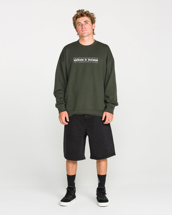 A young man wears the Volcom Men's Dirty Work Crew, a dark green crew sweatshirt crafted from recycled polyester with a graphic design, paired with black knee-length shorts, black socks, and black sneakers as he stands against a plain white background. The loose fit fleece provides both comfort and style for a laid-back, casual look.