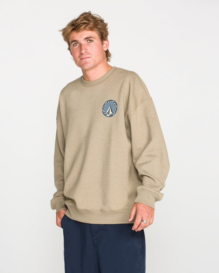 A person with short, light brown hair stands against a plain white background, wearing Volcom's Men's Dirty Work Crew—a beige, loose fit sweatshirt made from brushed-back fleece featuring a small round blue logo on the chest—paired with navy pants.
