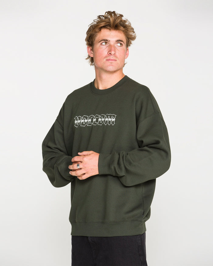 A person wearing the Volcom Men's Dirty Work Crew, a dark green crew sweatshirt with white graffiti-style text, stands against a plain background looking up and to the side with hands loosely clasped. The loose fit fleece—crafted from recycled polyester—adds comfort and style to their relaxed pose.