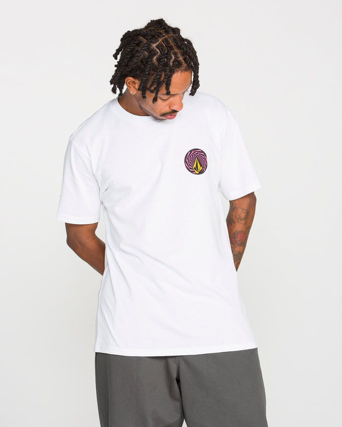A person with short twists and tattoos wears the Volcom Men's Dirty Work Short Sleeve Tee—a classic fit, white heavyweight cotton t-shirt featuring a small, colorful circular logo rendered in a soft-hand screen print on the left chest—paired with gray shorts, standing against a plain white background.