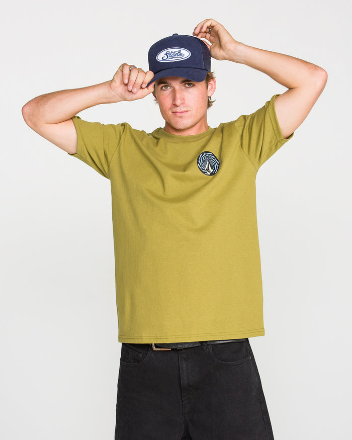 A person wearing a green Volcom Men's Dirty Work Short Sleeve Tee, paired with classic fit black pants and a navy baseball cap, adjusts their hat while looking at the camera against a plain white background.