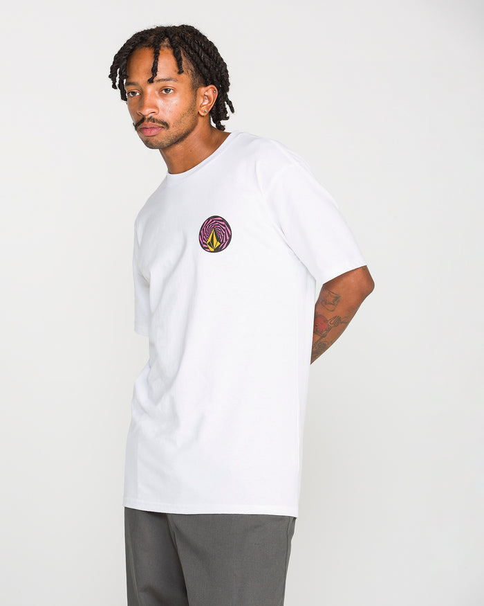 A man with short dreadlocks wears the Volcom Men's Dirty Work Short Sleeve Tee, crafted from heavyweight cotton and featuring a colorful circular logo on the chest in a soft-hand screen print. He pairs it with gray pants while standing against a plain, light background.