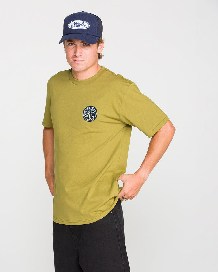 A person stands against a plain white background, hands on hips and looking at the camera, wearing a navy cap, black pants, and the Volcom Men's Dirty Work Short Sleeve Tee in olive green—a classic fit T-shirt featuring a circular logo on the chest. Crafted from premium ring-spun cotton, this tee offers both comfort and style.